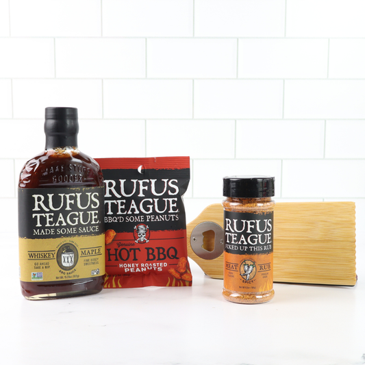 Three bottles of Rufus Teague sauce and a bag of peanuts on a white tiled background