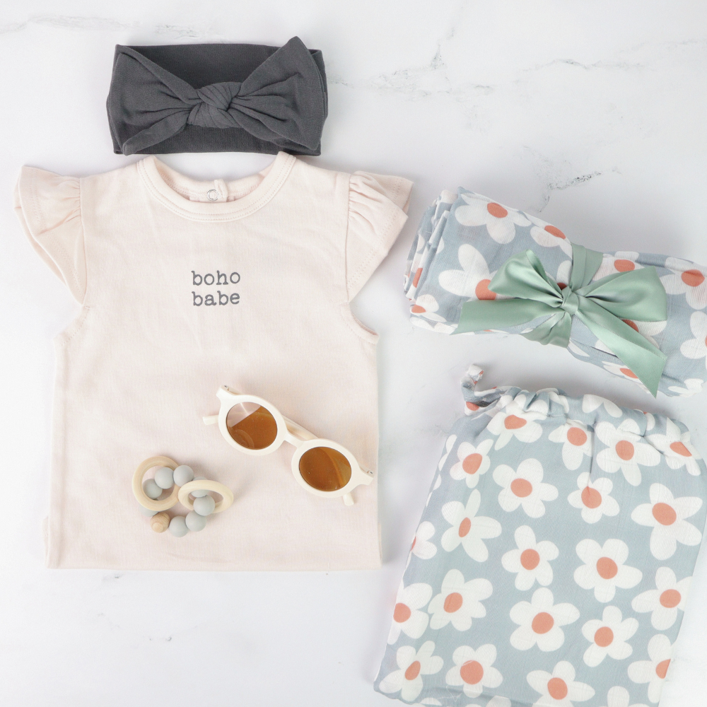 Baby clothing and accessories including a t-shirt, headband, sunglasses, and floral blanket on a light background.